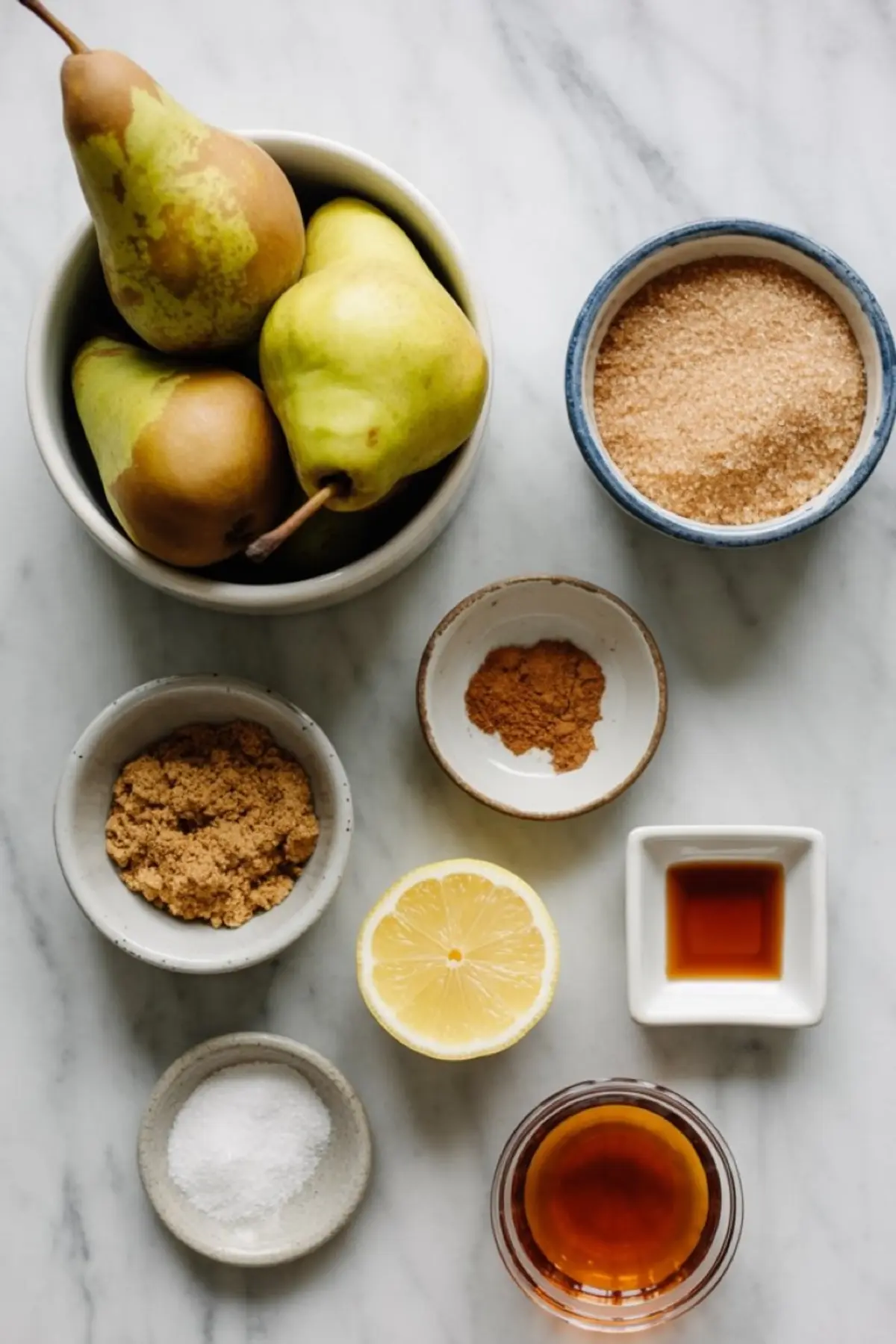 Flat lay of fresh pear butter ingredients on a marble surface, including green Bartlett pears, light and dark brown sugar, lemon, cinnamon, vanilla extract, salt, and apple cider.