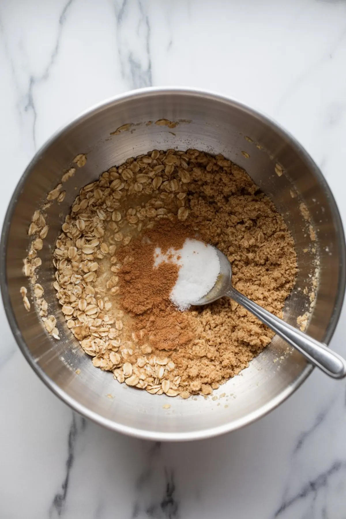 Stainless steel mixing bowl containing rolled oats, brown sugar, melted butter, and a spoonful of ground cinnamon and salt. The ingredients are partially combined for a crumble topping in a pear crisp recipe.
