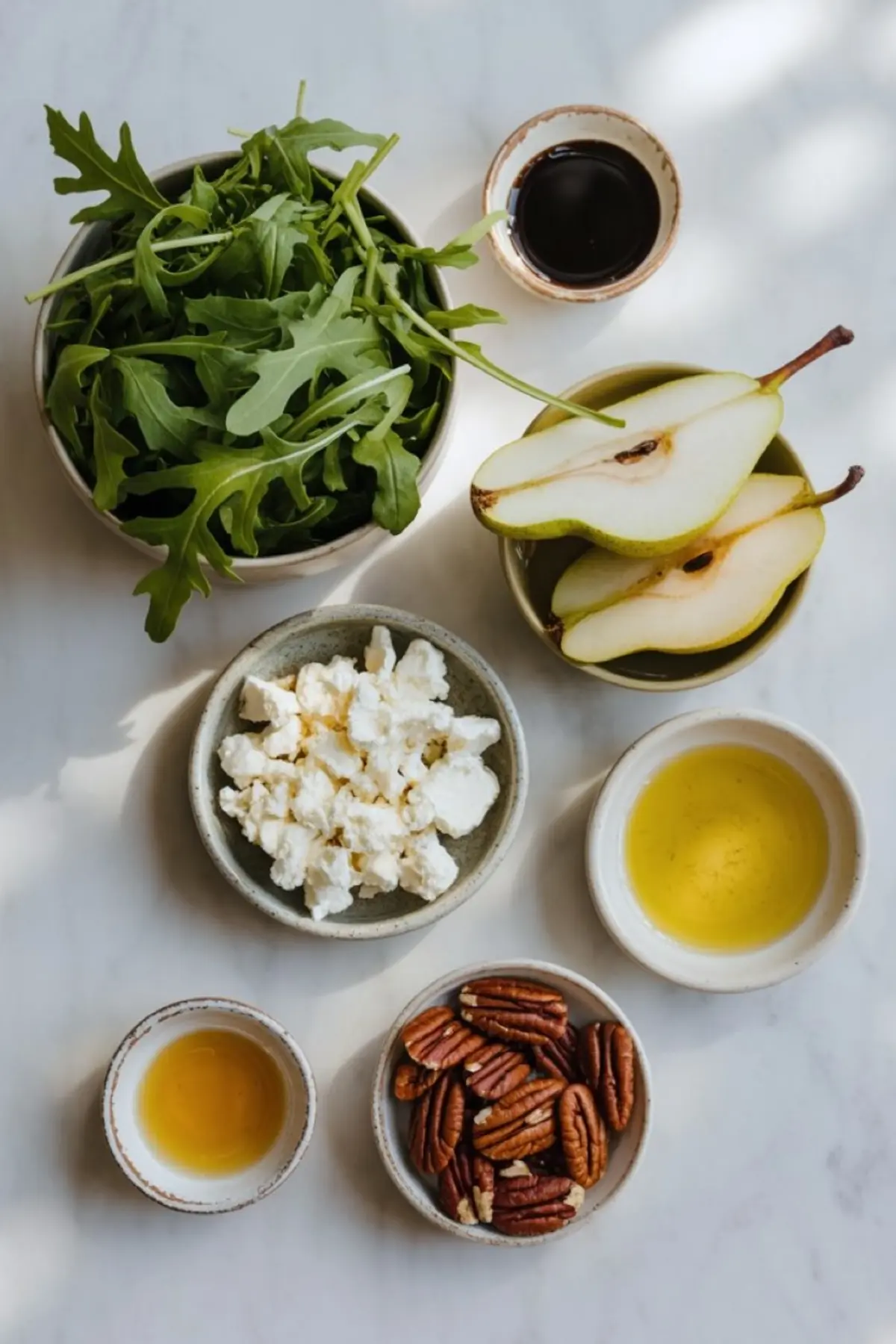 Overhead view of pear salad ingredients on a marble surface, including fresh arugula, halved pears, crumbled goat cheese, pecans, olive oil, balsamic vinegar, and honey in ceramic bowls.