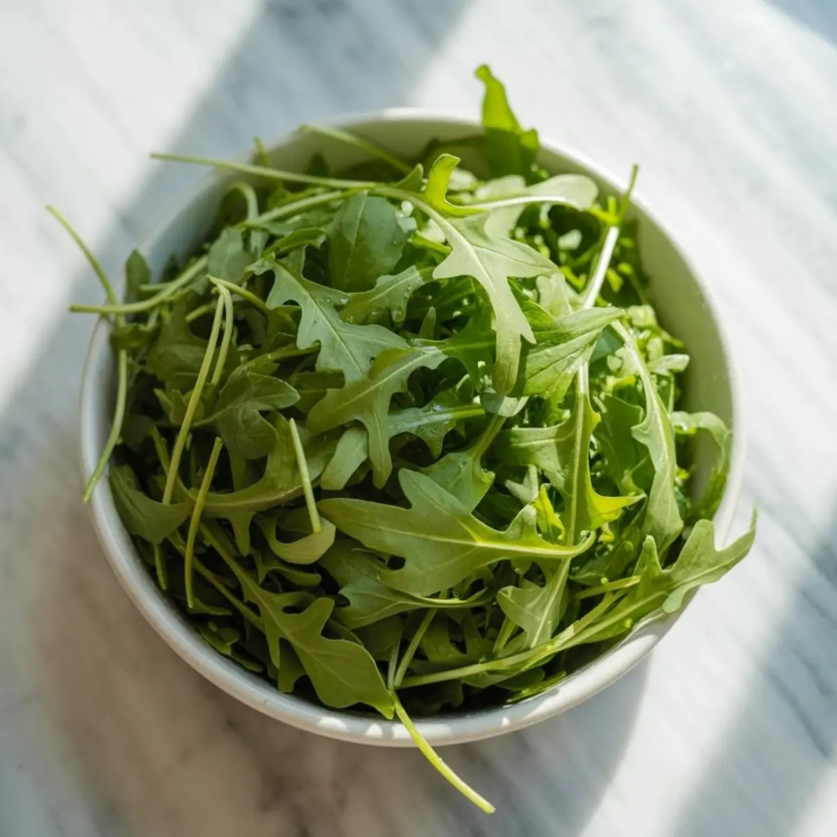 Close-up of a bowl filled with fresh arugula leaves on a marble countertop with natural light highlighting the greens.