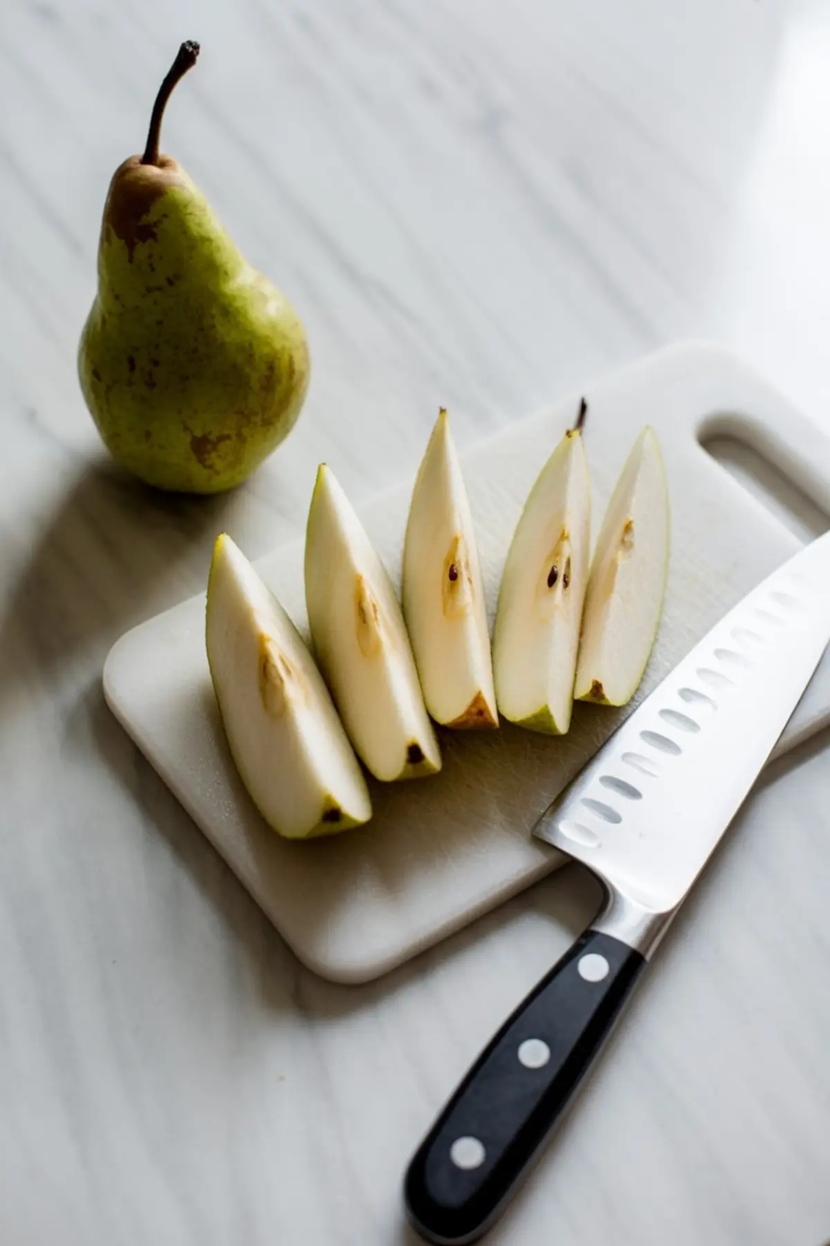 Sliced pear wedges arranged on a white cutting board next to a whole pear and a chef’s knife on a marble surface.