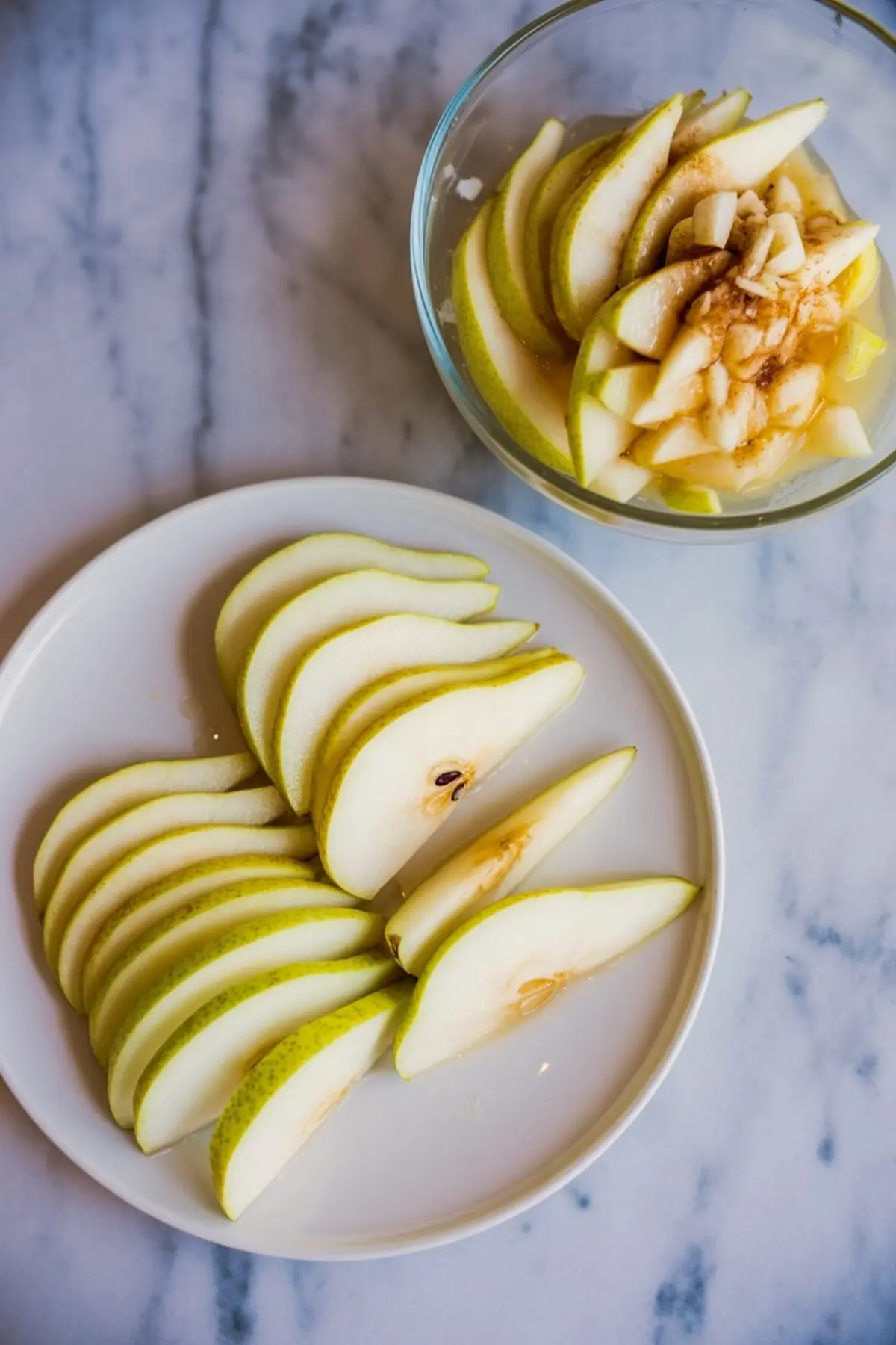 Sliced fresh pears arranged on a white plate and in a glass bowl, with the bowl showing the pears mixed with sugar and spices.
