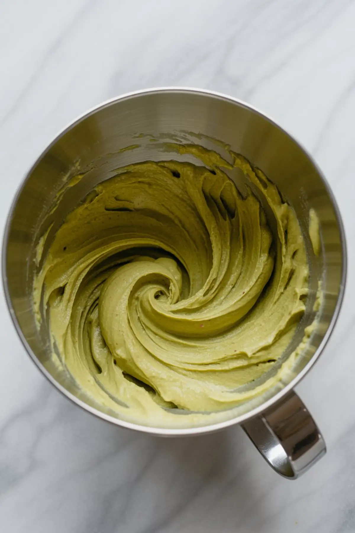 Swirled pistachio butter mixture inside a stainless steel mixing bowl on a marble surface, showcasing the creamy texture and vibrant green color of blended pistachios.