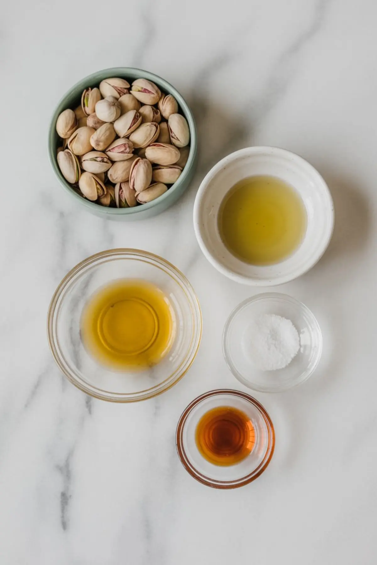Overhead view of pistachio butter ingredients on a white marble background, including shelled pistachios, olive oil, avocado oil, salt, and vanilla extract in separate glass and ceramic bowls.