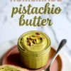 Labeled photo of homemade pistachio butter in a glass jar topped with chopped pistachios and served with a spoon and a slice of bread spread with the green nut butter on a pink plate.