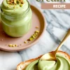A glass jar filled with silky pistachio cream is topped with chopped pistachios and placed on a pink plate, next to a slice of bread spread with the green cream and a gold spoon lifting a dollop.