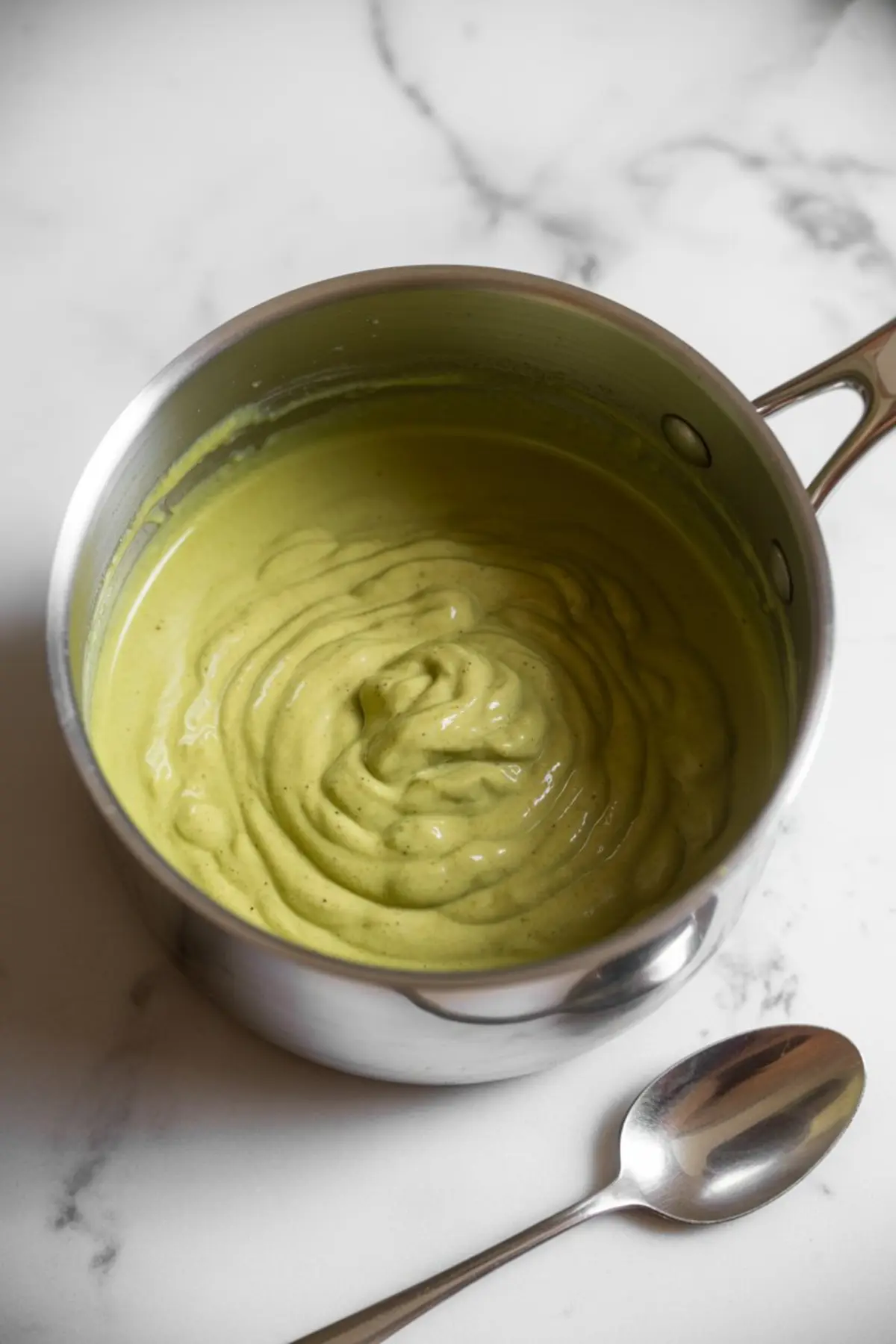 A stainless steel saucepan filled with smooth pistachio cream mixture sits on a marble counter with a silver spoon beside it, showing a creamy texture ideal for desserts or spreads.
