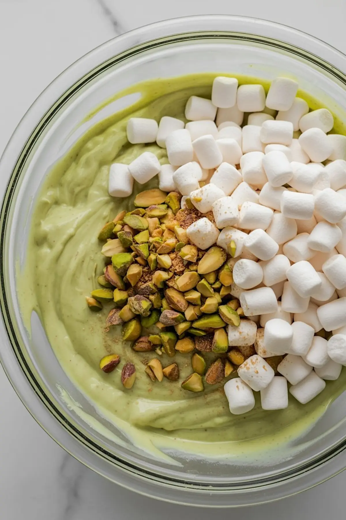 Overhead view of a mixing bowl filled with pistachio pudding, mini marshmallows, chopped pistachios, and brown sugar before mixing.