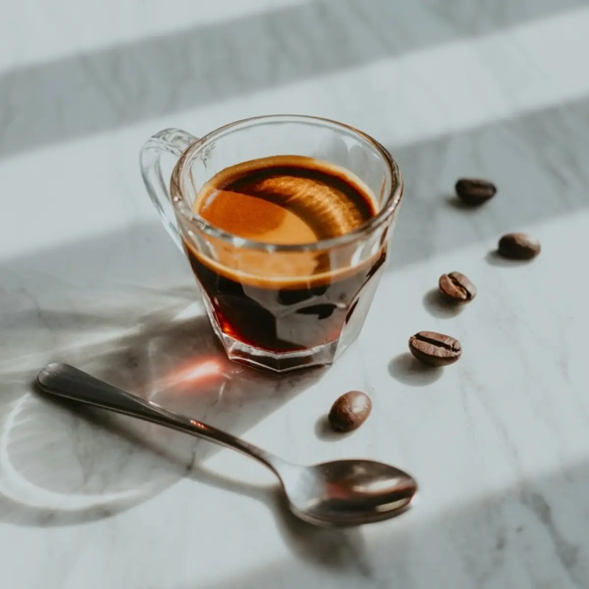 Glass of freshly brewed espresso with rich crema on a marble surface, accompanied by whole coffee beans and a stainless steel spoon in natural light.
