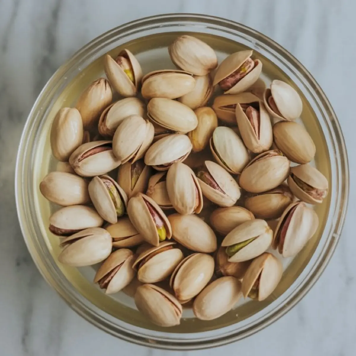 Top view of a bowl filled with raw pistachios soaking in water, placed on a white marble countertop.

