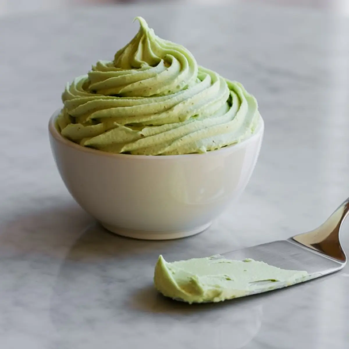 Small white bowl of light green pistachio buttercream frosting piped in a swirl pattern, with a metal spatula showcasing a smooth sample on marble countertop.