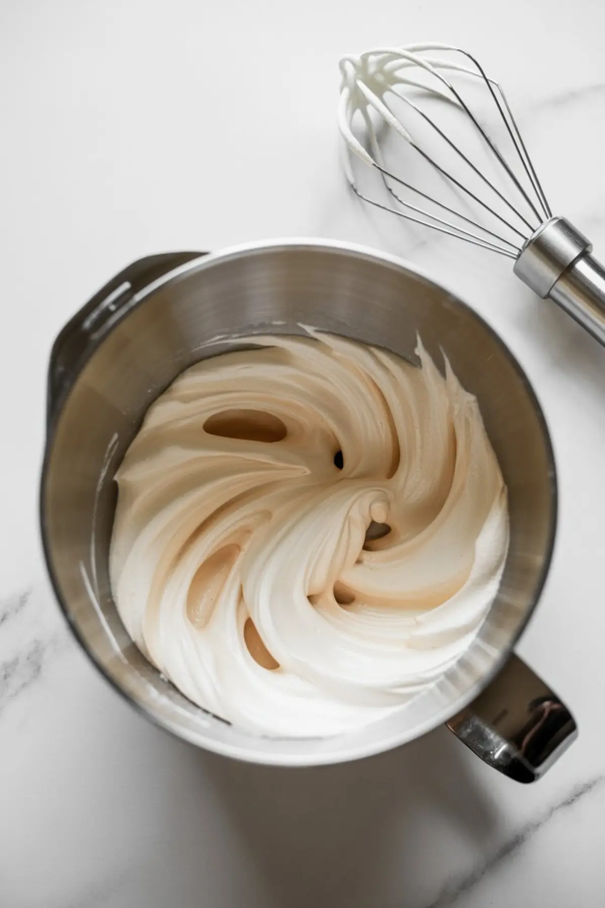 Mixing bowl of freshly whipped glossy meringue with soft peaks, beside a metal balloon whisk coated with the mixture on white marble background.