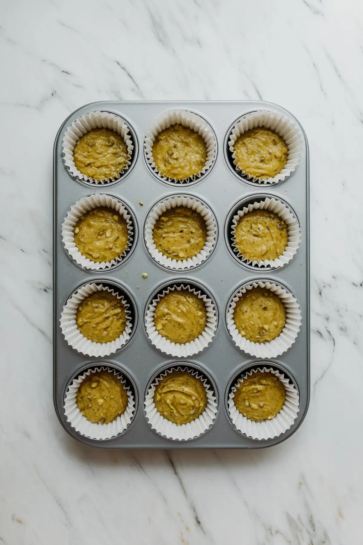Overhead shot of a muffin tray filled with unbaked pistachio muffin batter in white paper liners, ready for baking on a marble surface.