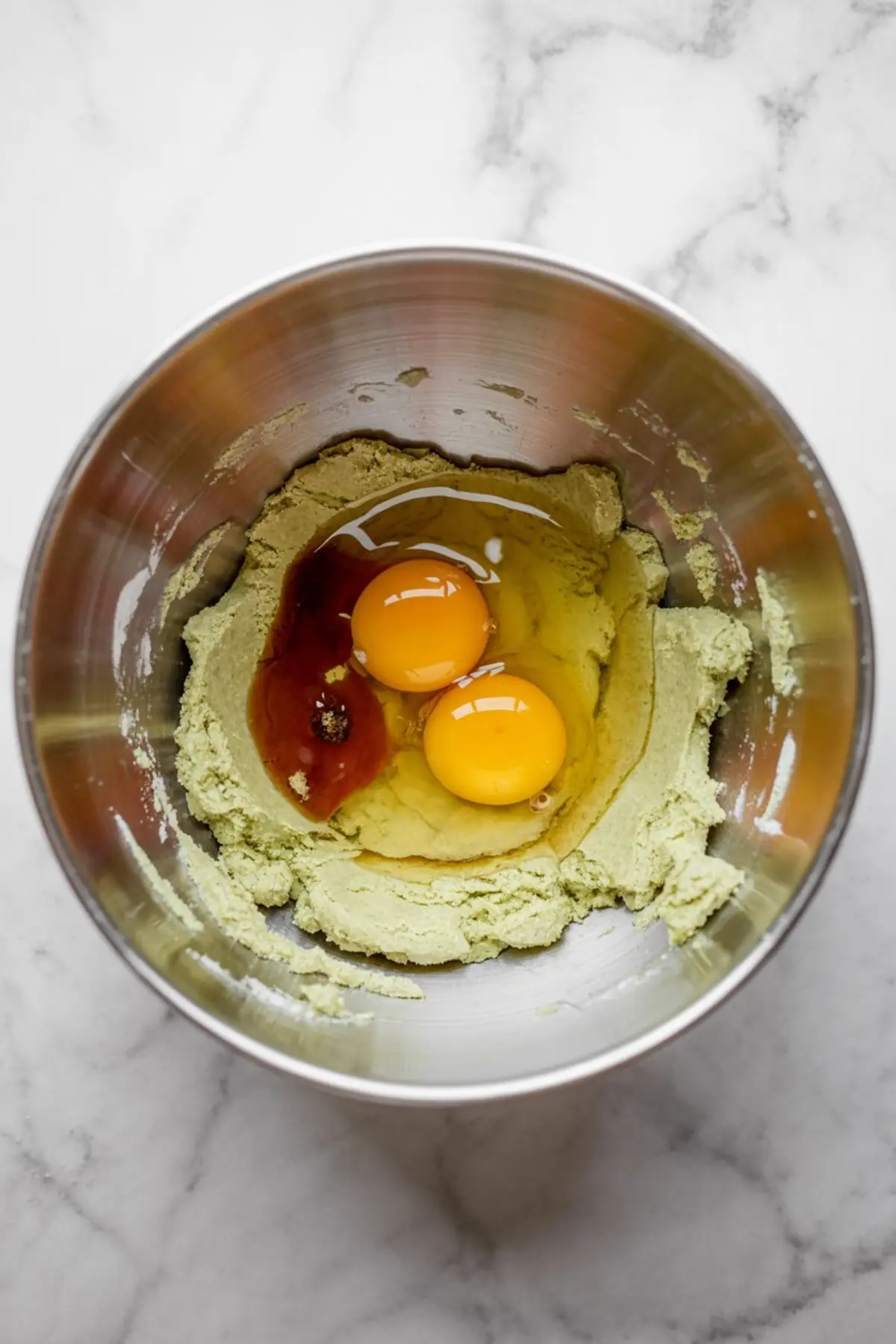 Mixing bowl filled with partially mixed pistachio cookie dough topped with raw eggs and vanilla extract, ready for blending.