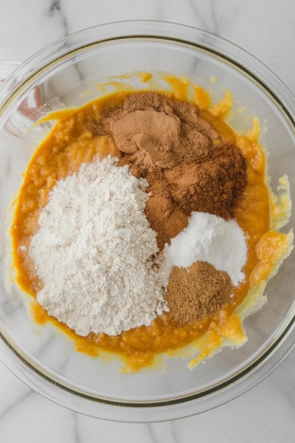 A mixing bowl of pumpkin batter topped with dry ingredients—flour, cinnamon, nutmeg, ginger, and baking soda—ready to be stirred.