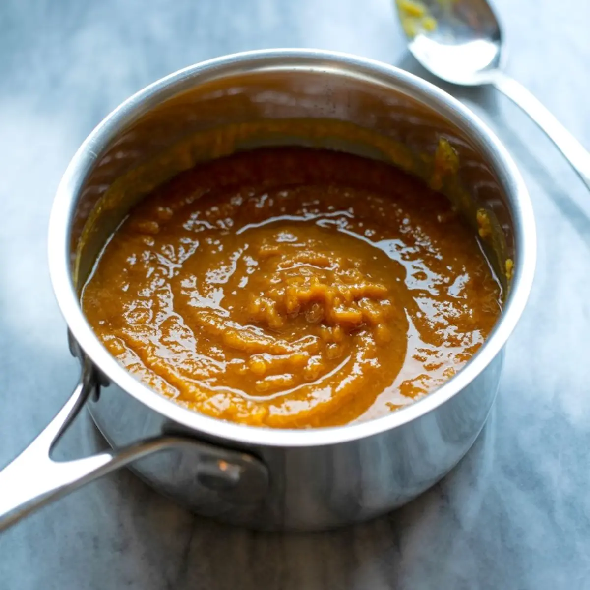 Smooth and glossy pumpkin butter swirled in a stainless steel saucepan, ready for cooling or jar filling on a marble background.