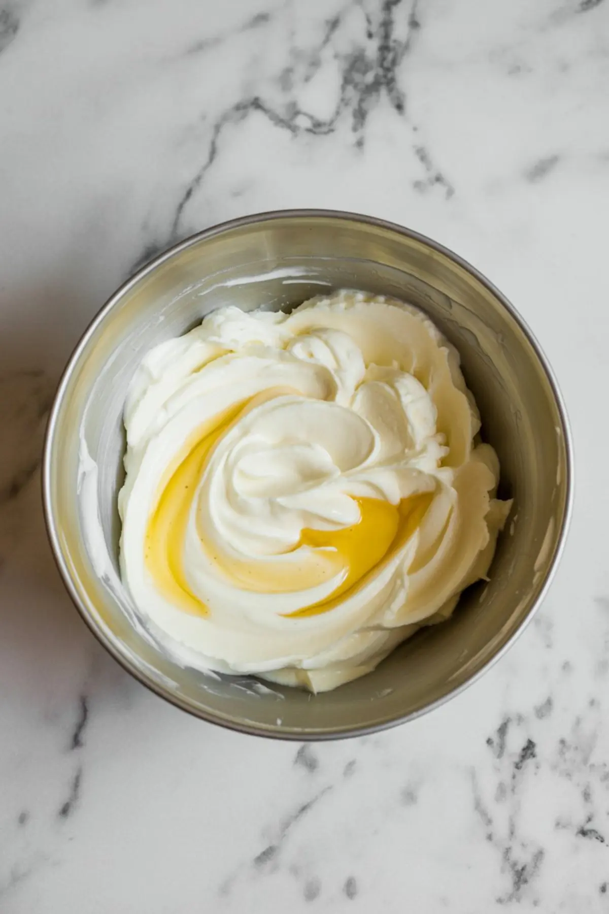 Fluffy whipped cream cheese mixture in a metal mixing bowl with golden swirls of vanilla extract on a marble countertop.