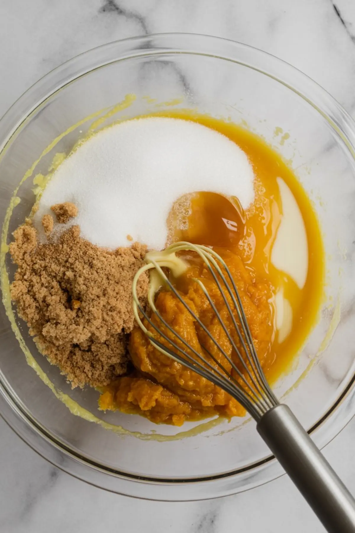 Brown sugar, granulated sugar, eggs, pumpkin purée, milk, vanilla, and oil in a glass mixing bowl with a metal whisk, combining to form the base for moist pumpkin choc chip muffins.
