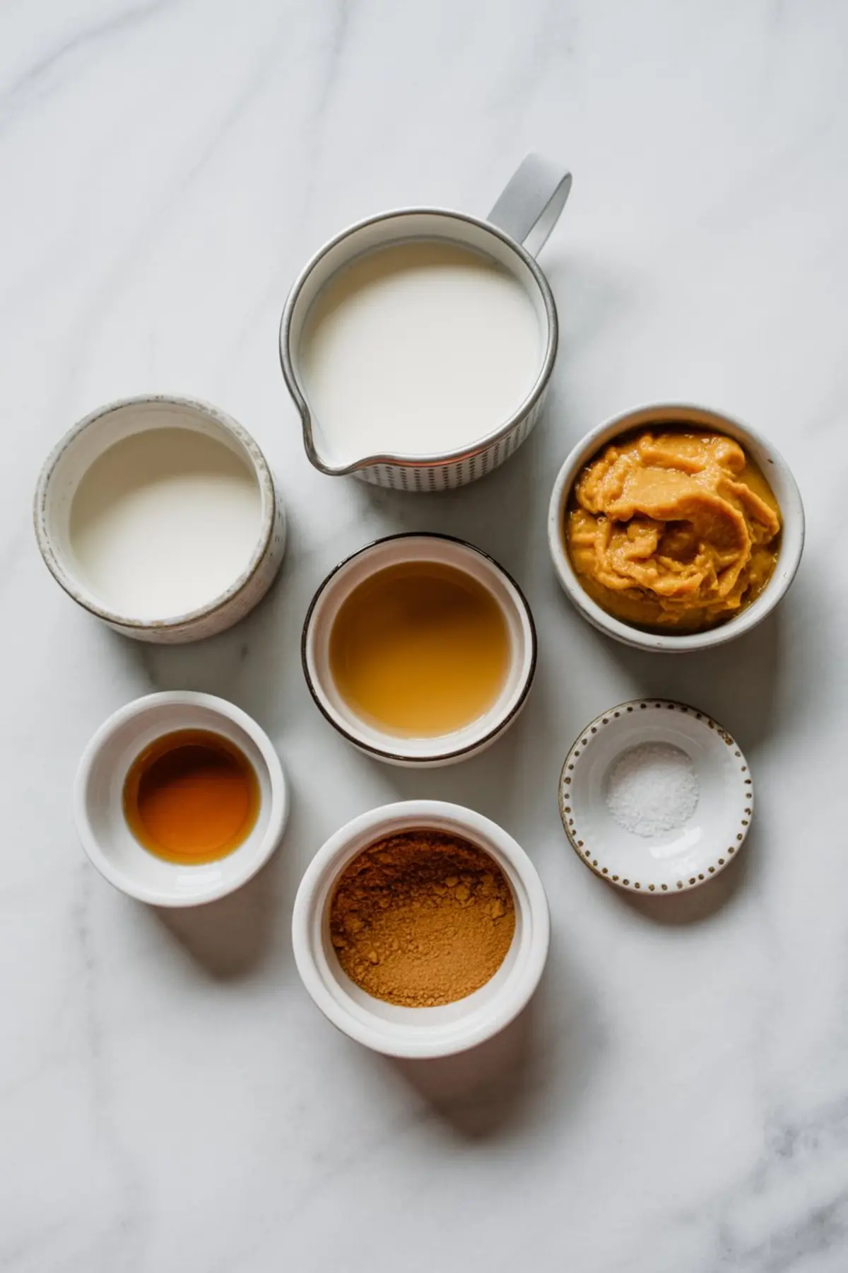 Overhead view of small bowls with ingredients for pumpkin coffee creamer, including milk, pumpkin puree, maple syrup, vanilla extract, cinnamon, and salt.