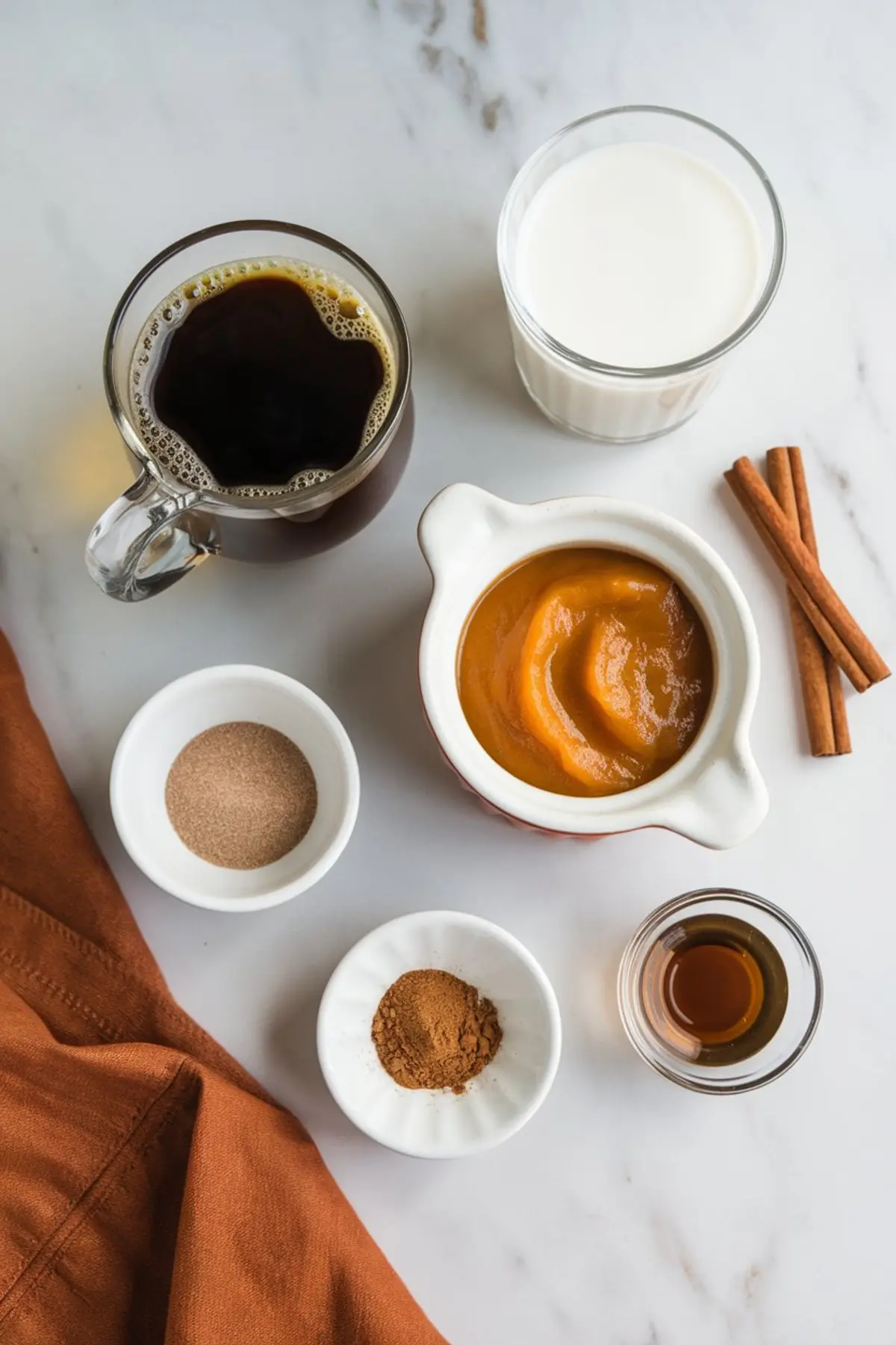 Flat lay of pumpkin spice coffee ingredients including brewed coffee, milk, pumpkin puree, ground cinnamon, pumpkin pie spice, vanilla extract, and sugar on a marble countertop with cinnamon sticks and a brown napkin.