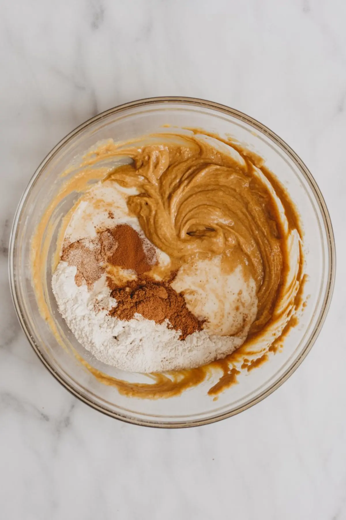 Glass mixing bowl showing partially mixed pumpkin muffin batter with flour, cinnamon, and other spices on top.
