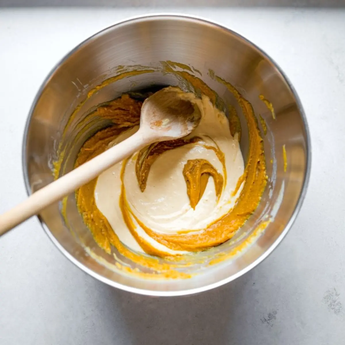 Metal mixing bowl with a wooden spoon stirring together pumpkin puree and cream cheese batter on a light gray countertop.