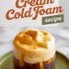 A glass of cold brew topped with thick pumpkin cream cold foam, garnished with a sprinkle of cinnamon, placed on a green ceramic plate with “Pumpkin Cream Cold Foam recipe” text above.
