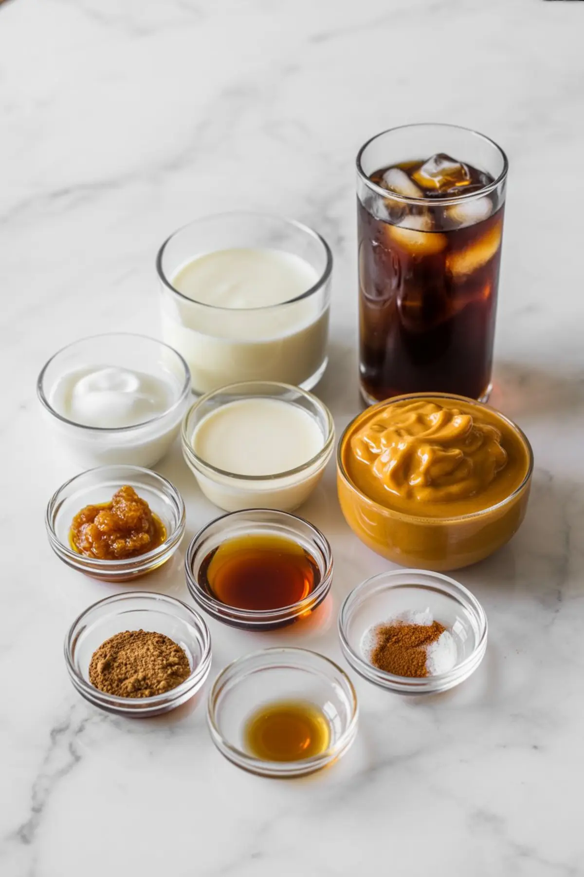 A flat lay of ingredients in clear glass bowls and cups arranged on a marble surface, featuring pumpkin purée, cold brew coffee, milk, yogurt, maple syrup, brown sugar syrup, vanilla extract, pumpkin spice, and cinnamon.