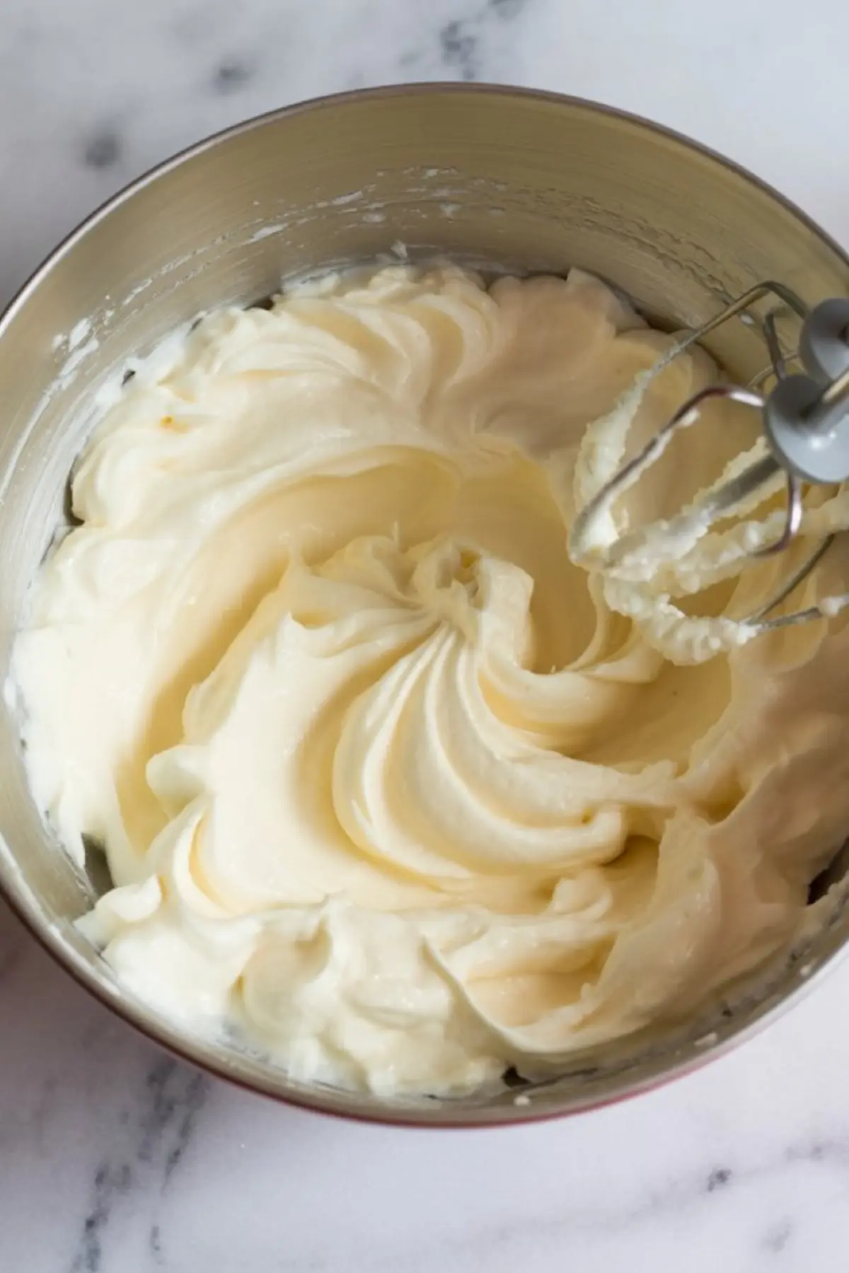 Whipped cream cheese mixture in a metal bowl with electric mixer beaters showing light, fluffy texture on a marble countertop.