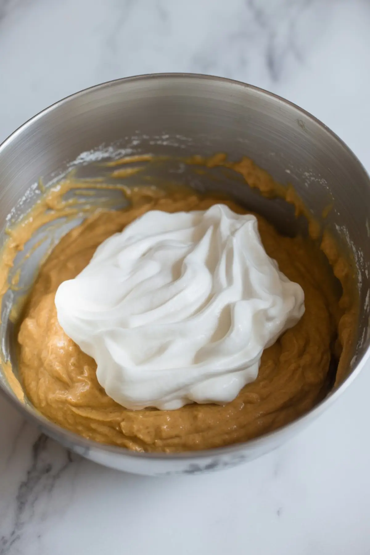 Metal bowl filled with pumpkin mixture topped with whipped cream, ready to be folded in, placed on a white marble counter.