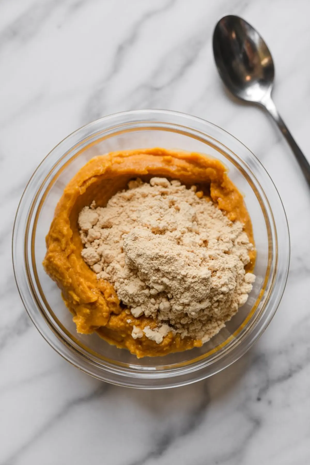 Clear glass bowl filled with pumpkin puree topped with vanilla protein powder on a marble countertop, with a metal spoon beside it, showcasing ingredients before blending for a pumpkin protein fluff.