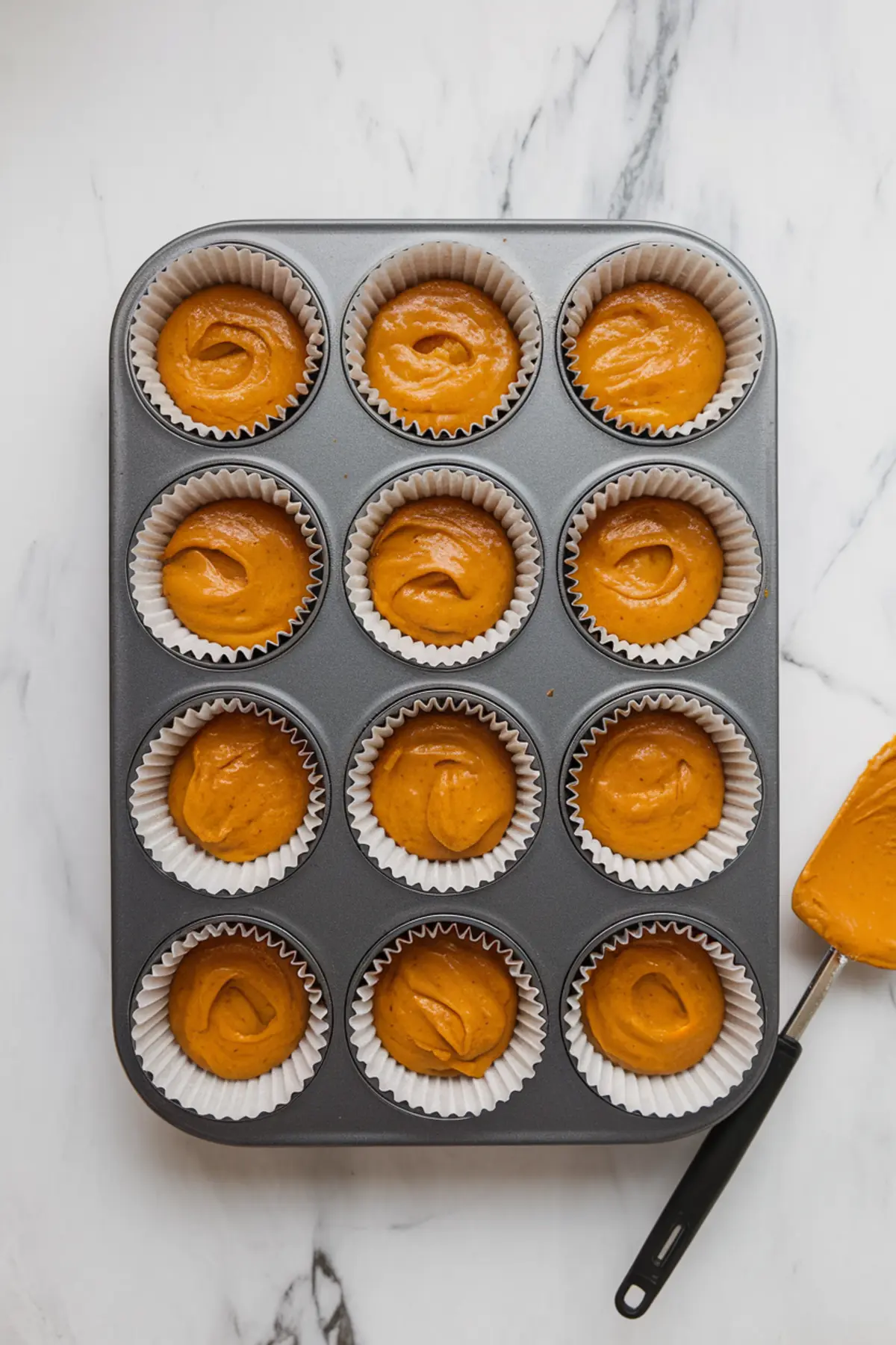 Pumpkin batter scooped into white paper liners inside a metal 12-cup muffin pan, arranged on a marble countertop ready for baking.