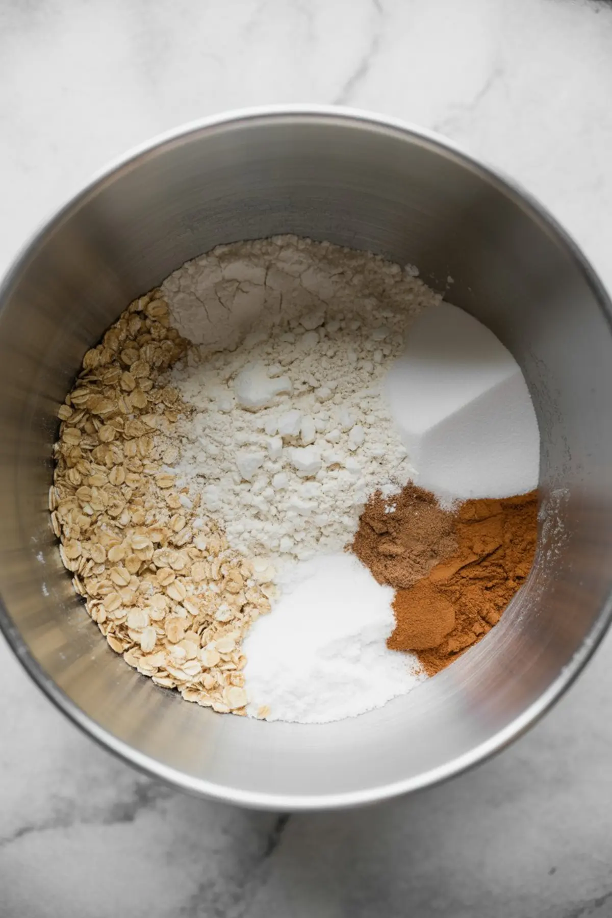 Overhead view of dry ingredients in a stainless steel mixing bowl, including rolled oats, flour, baking soda, baking powder, salt, granulated sugar, ground cinnamon, and ground nutmeg.