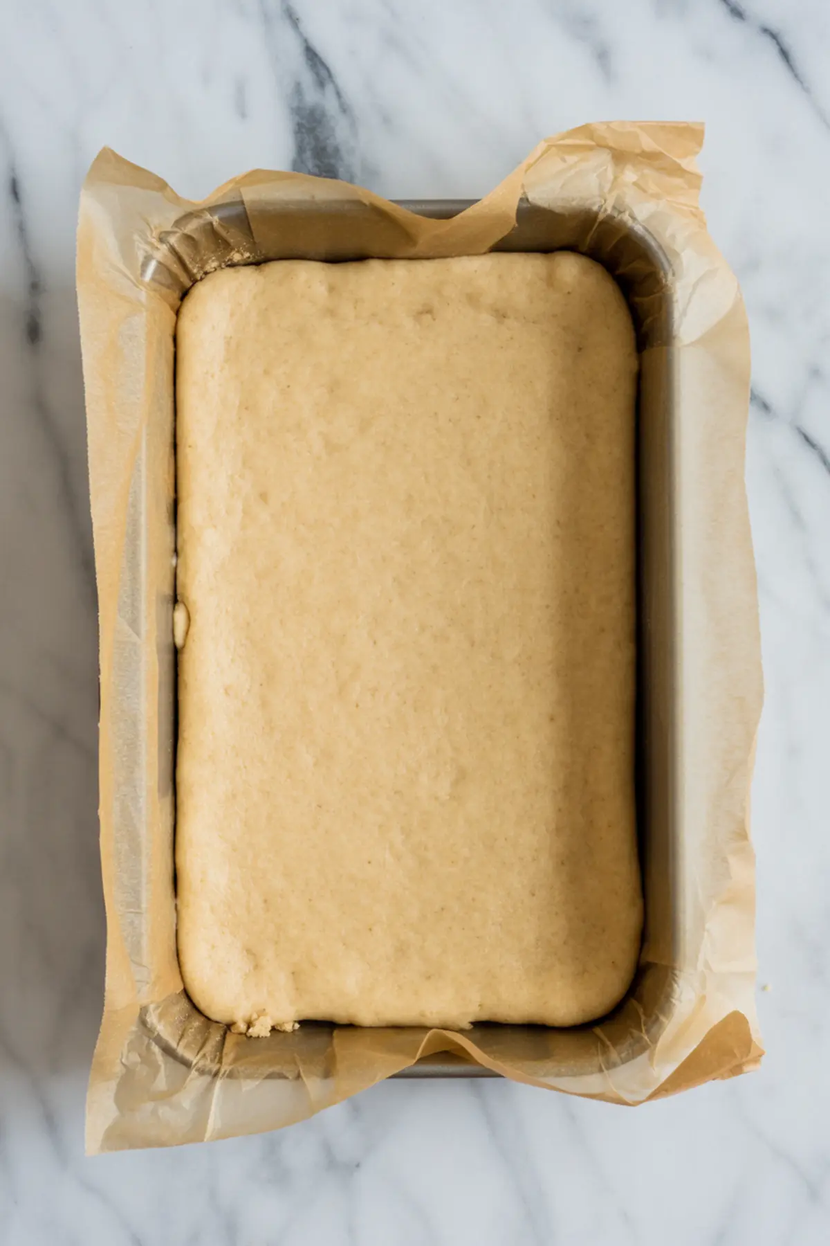 Rectangular baking pan lined with parchment paper filled with an even layer of raw crust dough, placed on a marble surface.