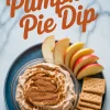 Overhead view of pumpkin pie dip garnished with cinnamon in a glass bowl, served with fresh apple slices and stacked graham crackers on a blue plate, with “Pumpkin Pie Dip” text in bold at top.