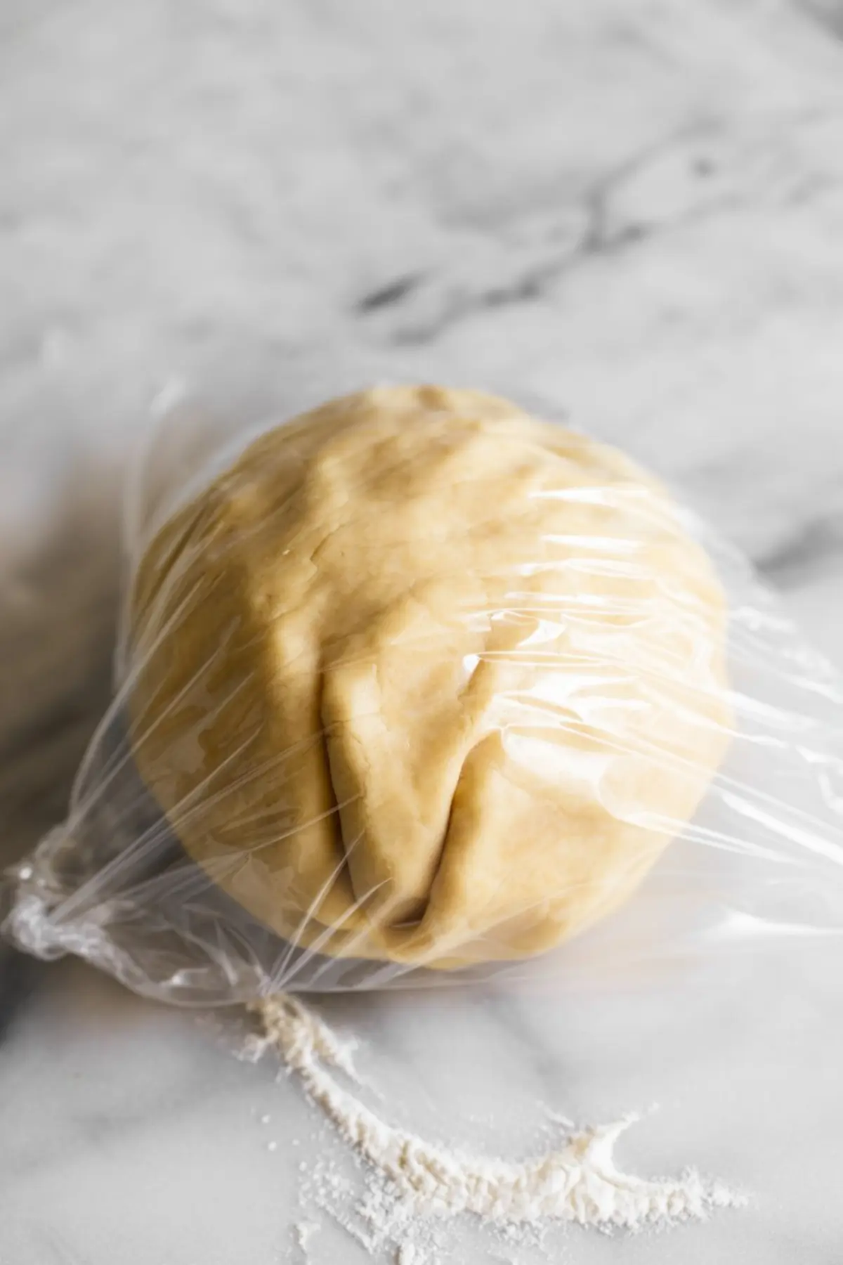 Ball of pie dough wrapped in plastic wrap on a floured marble surface, ready for chilling before rolling.
