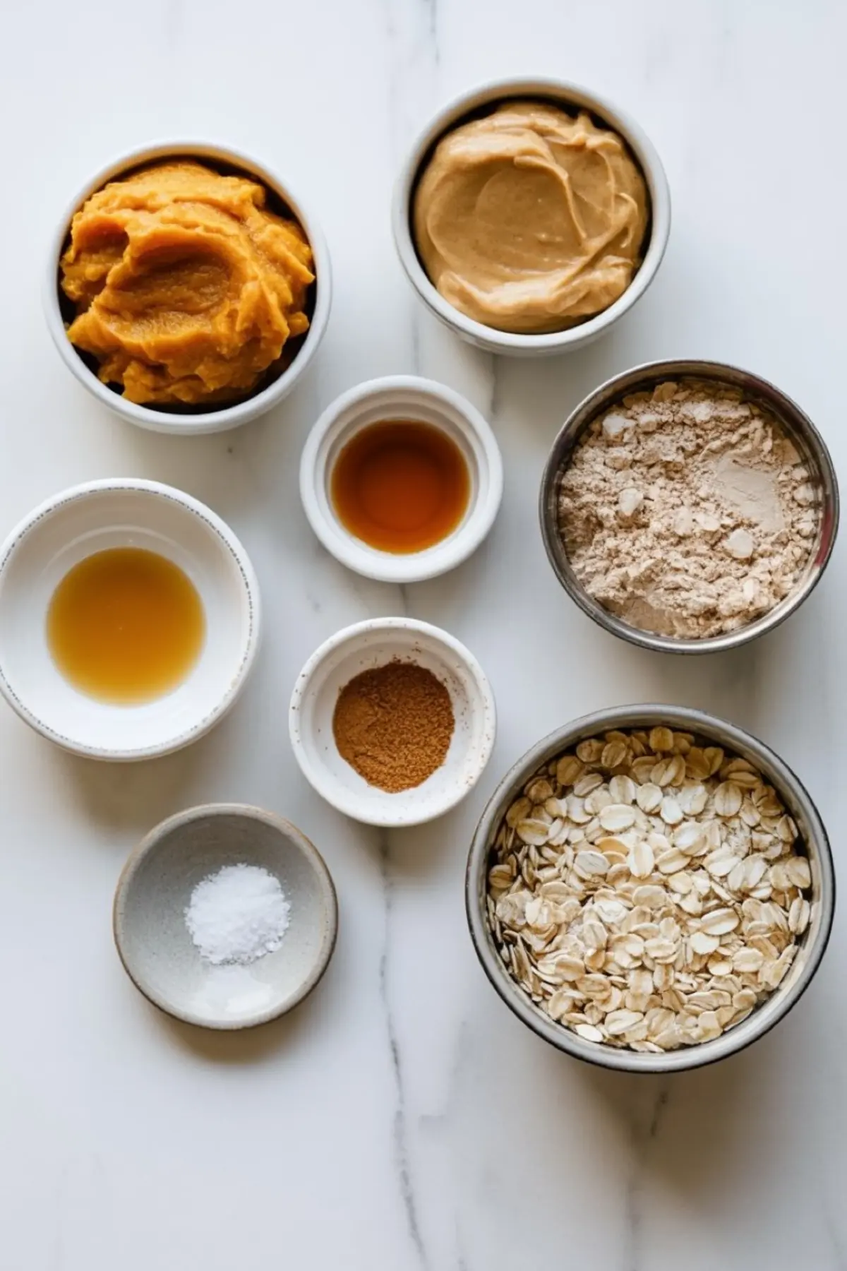 Flat lay of pumpkin protein ball ingredients on white marble, including pumpkin puree, peanut butter, vanilla extract, maple syrup, cinnamon, oat flour, rolled oats, and sea salt.