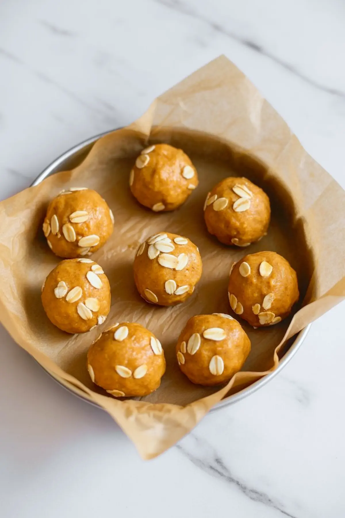Round baking tin lined with parchment paper holding seven pumpkin protein balls, each topped with rolled oats, ready for chilling or baking.