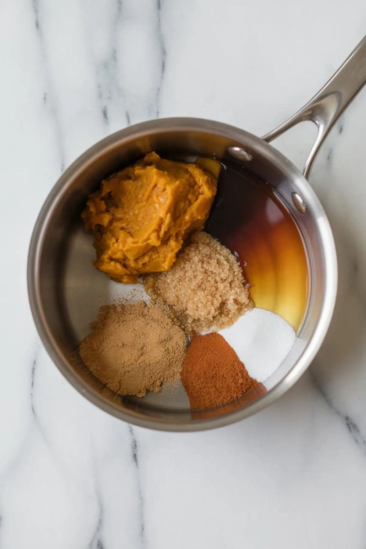 Overhead view of a stainless steel saucepan filled with pumpkin puree, brown sugar, granulated sugar, maple syrup, and warm spices like cinnamon, ginger, and nutmeg, set on a marble surface.