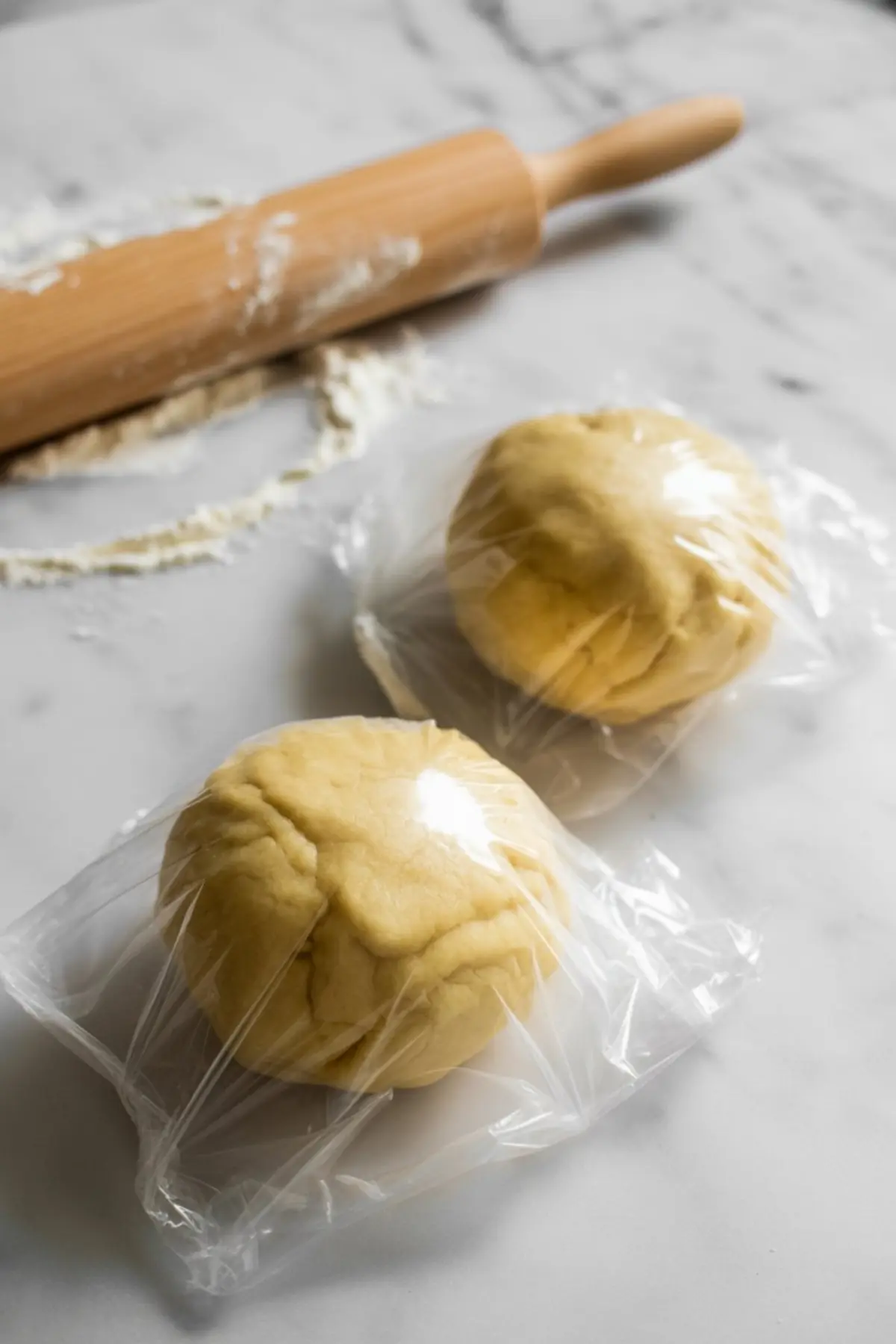 Two portions of cookie dough wrapped in plastic wrap resting on a floured marble countertop beside a wooden rolling pin—chilled dough ready for rolling.