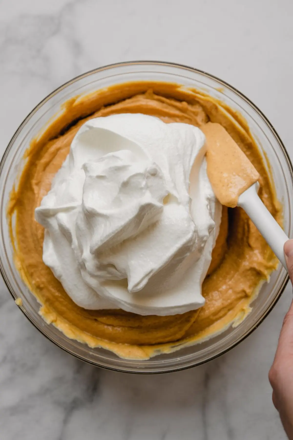 Large bowl of pumpkin and cream mixture with whipped topping being folded in using a spatula, showing the smooth texture of the filling.