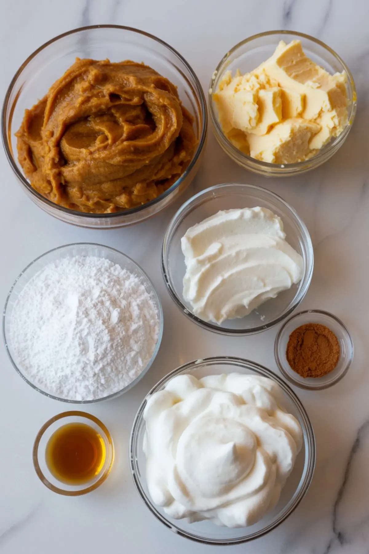Assorted ingredients in glass bowls on a white surface, including pumpkin puree, softened butter, whipped topping, powdered sugar, vanilla extract, cream cheese, and pumpkin spice.