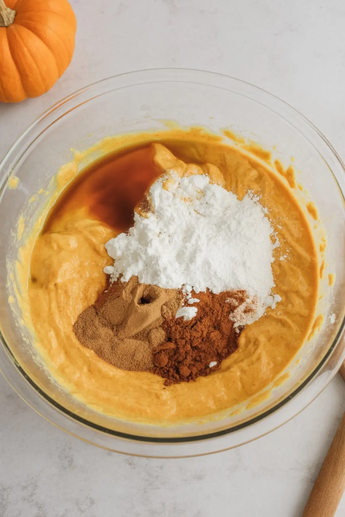 Glass mixing bowl filled with pumpkin puree batter, topped with powdered sugar, vanilla extract, ground cinnamon, and ground ginger on a light countertop with a small pumpkin in the corner.