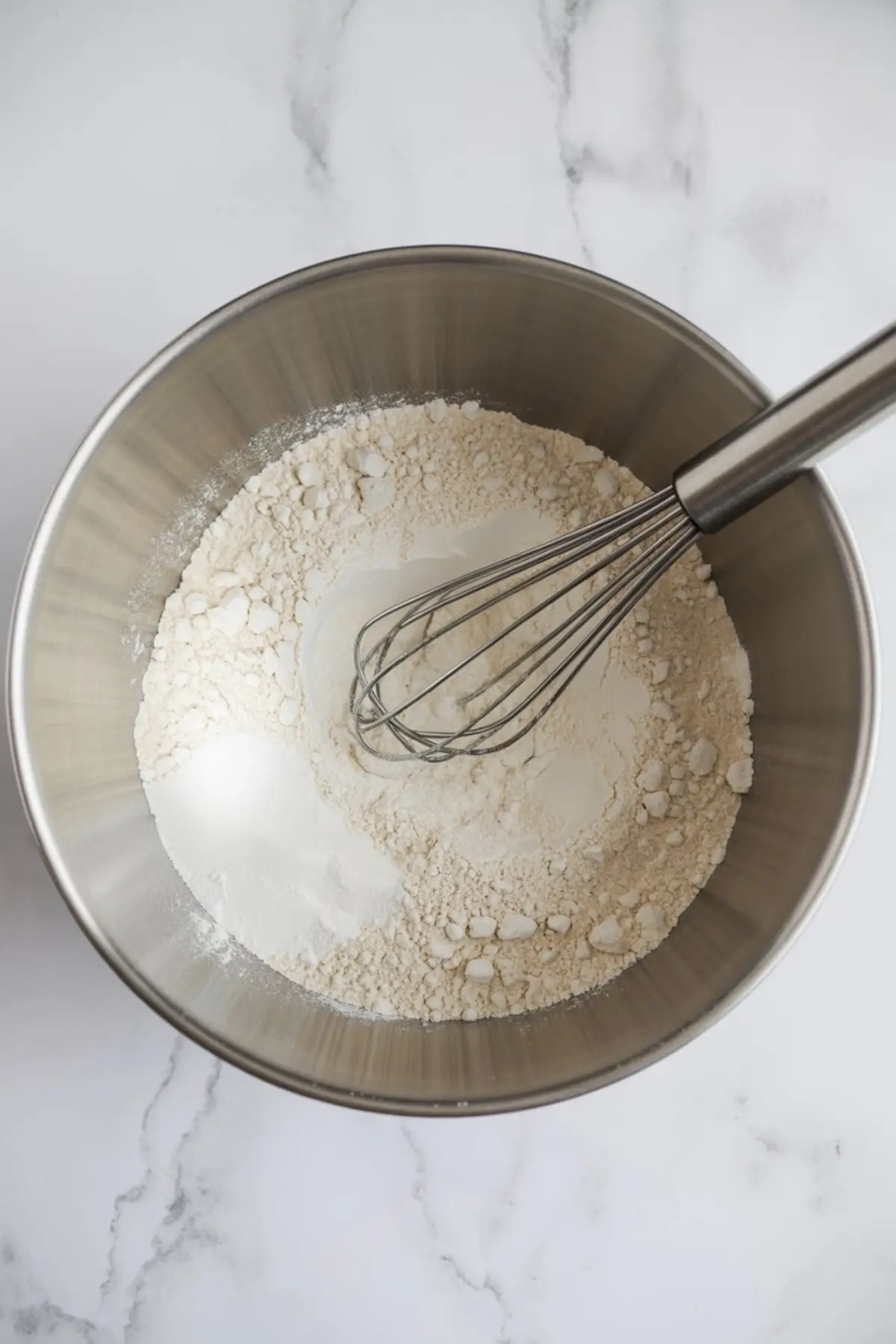 Large mixing bowl with flour, sugar, and baking powder being whisked together on a marble counter.