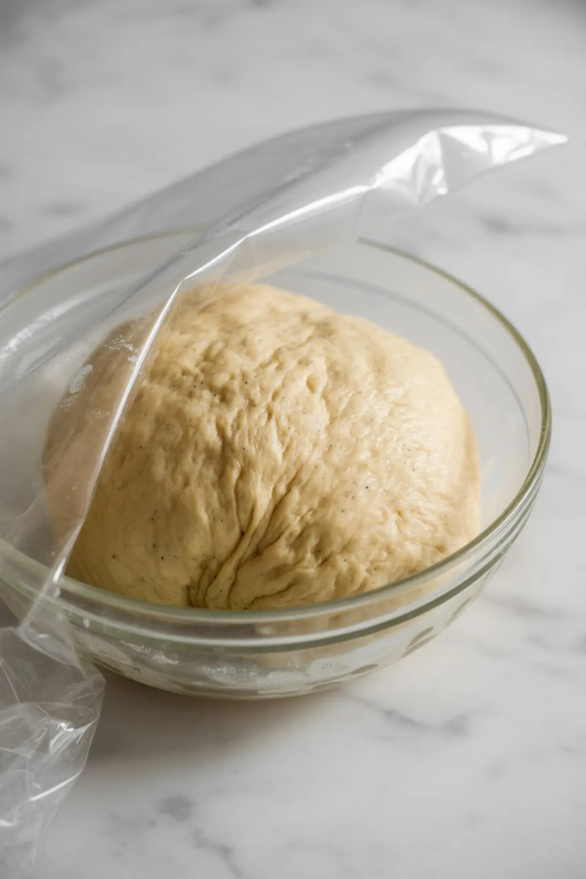Glass bowl with risen dough partially covered with plastic wrap.