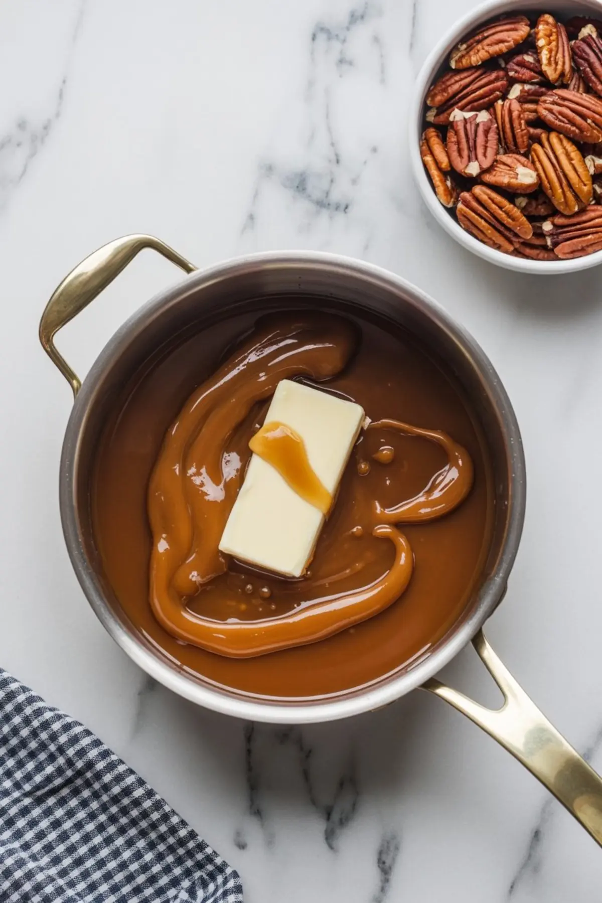 Saucepan with melted caramel and butter, placed next to a bowl of pecans on a marble surface.