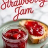 Jar of strawberry jam with visible fruit pieces on a plate beside a slice of bread spread with jam and a spoonful resting on the side, text overlay reading “Strawberry Jam.”