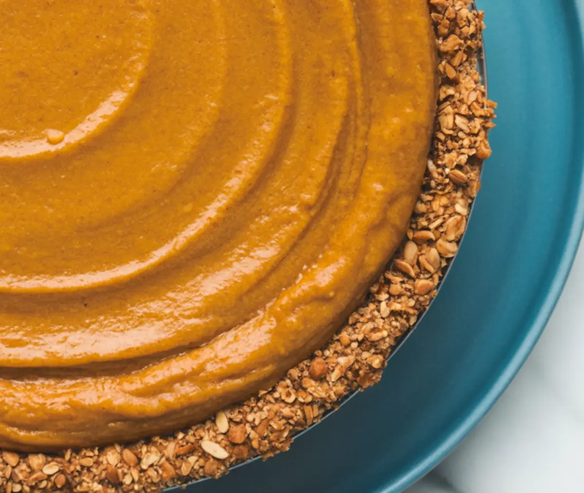 Close-up of a vegan pumpkin pie on a blue plate, highlighting the glossy swirl of the pumpkin filling and textured oat and nut crust.
