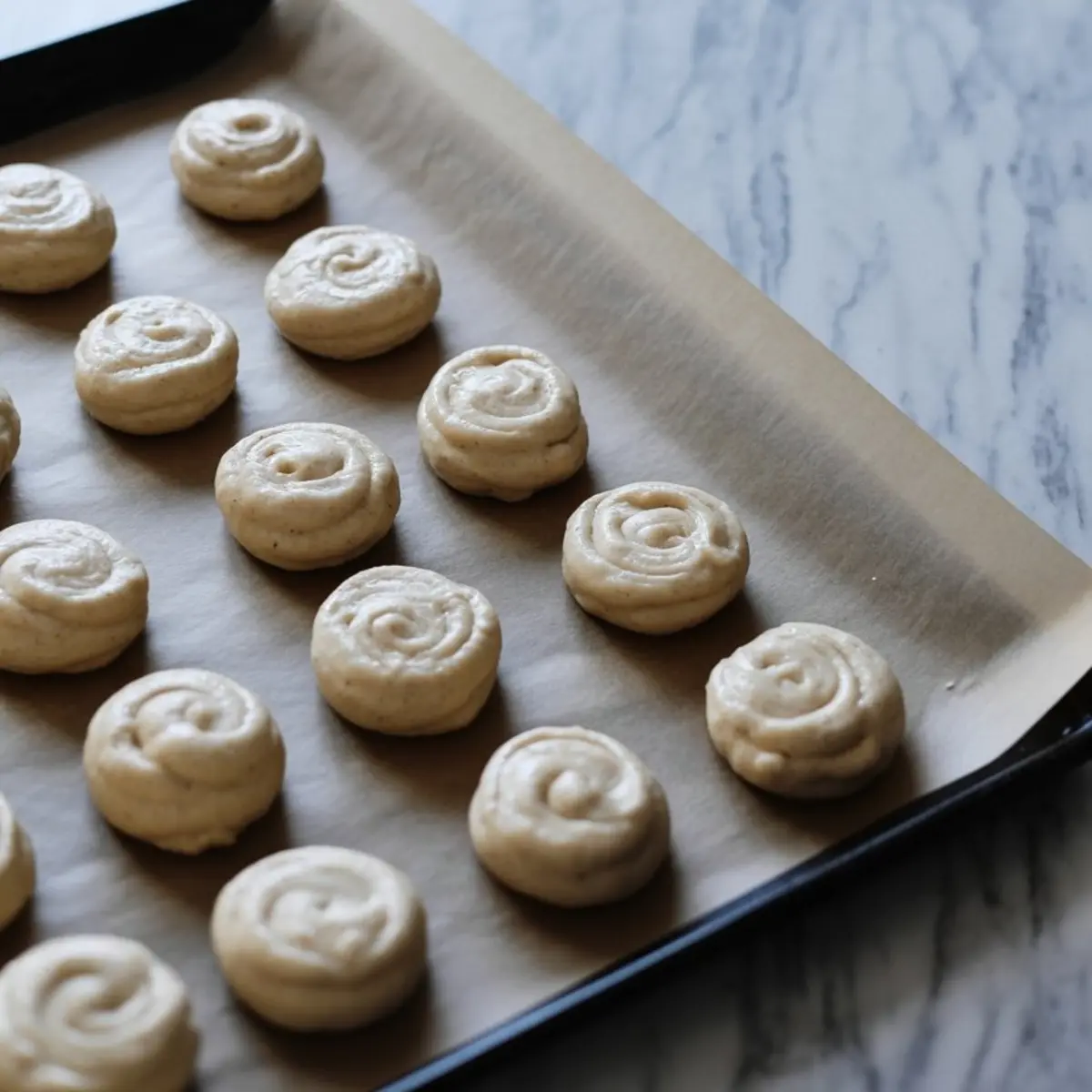 Piped cookie dough rounds with swirled tops arranged in rows on a parchment-lined baking sheet, ready for baking.