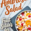 Flat lay of a serving bowl filled with ambrosia salad placed on a gray plate over a marble surface, surrounded by mini marshmallows and featuring text “Ambrosia Salad – Fruit Salads for Parties.”